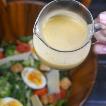 Homemade Caesar Salad Dressing in a small seethrough pitcher held over a wooden salad bowl full of greens, hard boiled egg halves and tomatoes