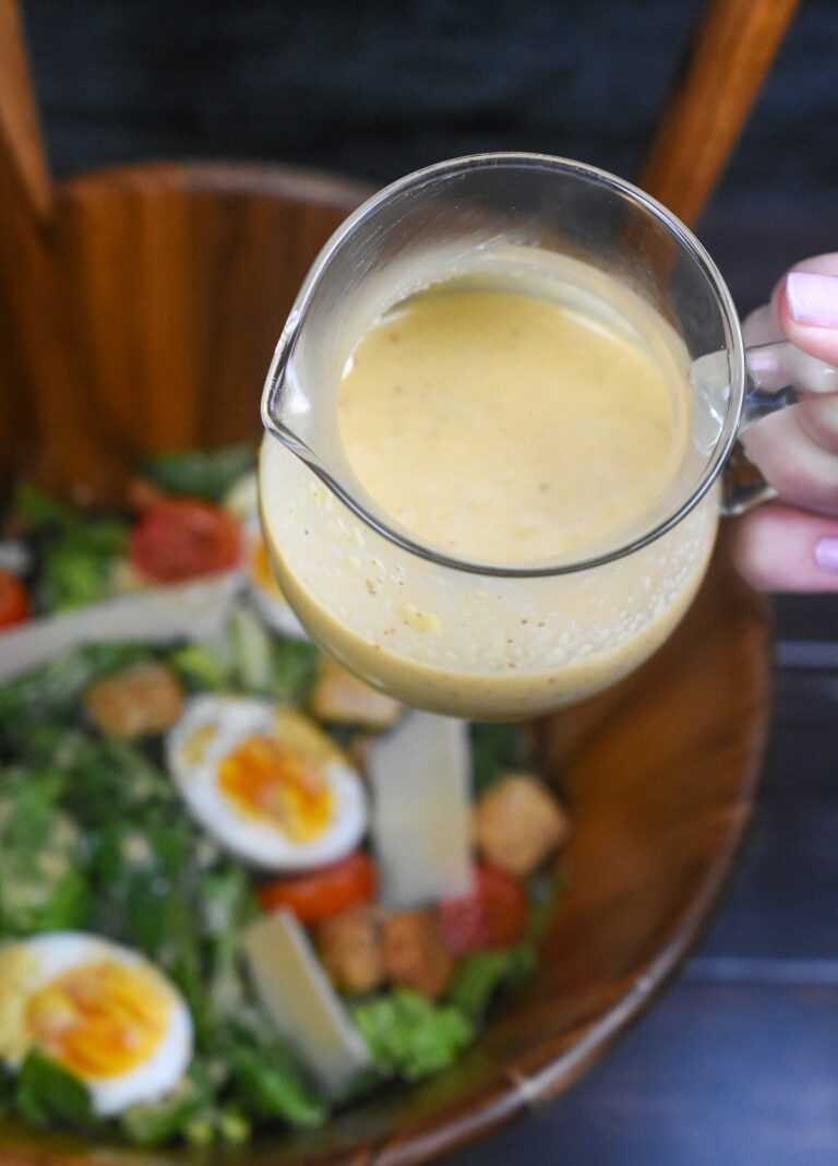 Homemade Caesar Salad Dressing in a small seethrough pitcher held over a wooden salad bowl full of greens, hard boiled egg halves and tomatoes