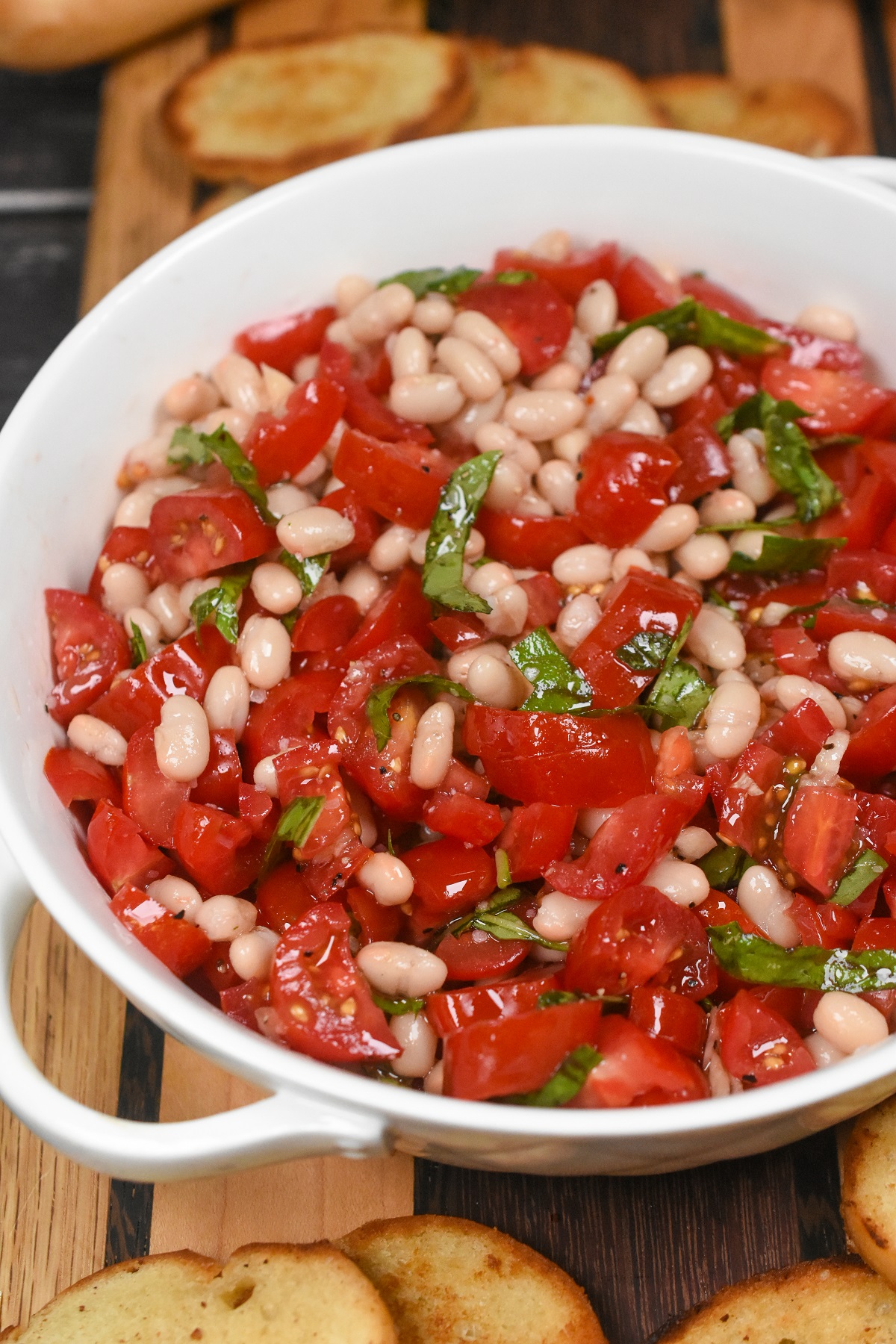 White Bean and Tomato Bruschetta. White Bean Dip with tomatoes. Shown small white beans with chopped tomatoes and ribbons of fresh basil in a white dish tossed with seasoned garlic oil. Crostini rounds in background.