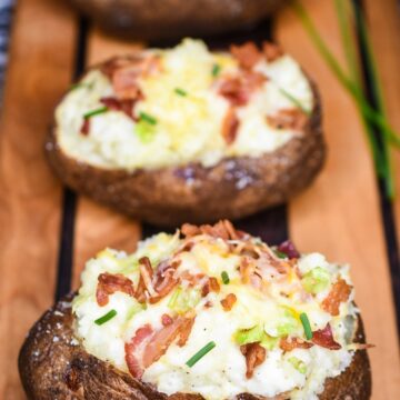 How to make Twice Baked Potatoes. Shown 3 twice baked potatoes topped with bacon cheese and chives on a wood board