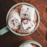 Hot cocoa in a pale green ceramic mug topped with marshmallows and cocoa powder
