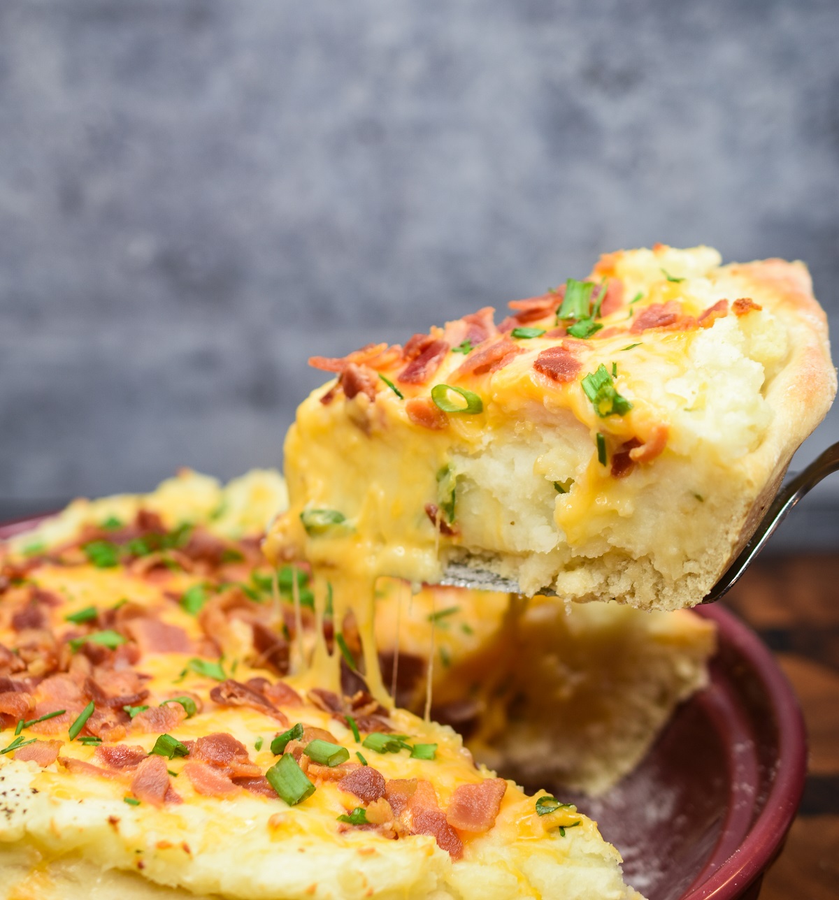 Mashed Potato Pizza topped with cheese, bacon and chives. Shown one thick and deep slice being lifted out of pie plate with cheese pull.