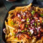 CAst iron skillet filled with French fries topped with pulled pork with BBQ sauce drizzle, scallions and red onion
