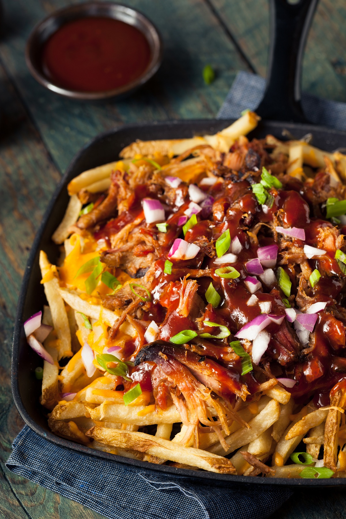 Cast iron pan with French fries topped with pulled pork. Garnished with barbecue sauce, scallions and red onions.