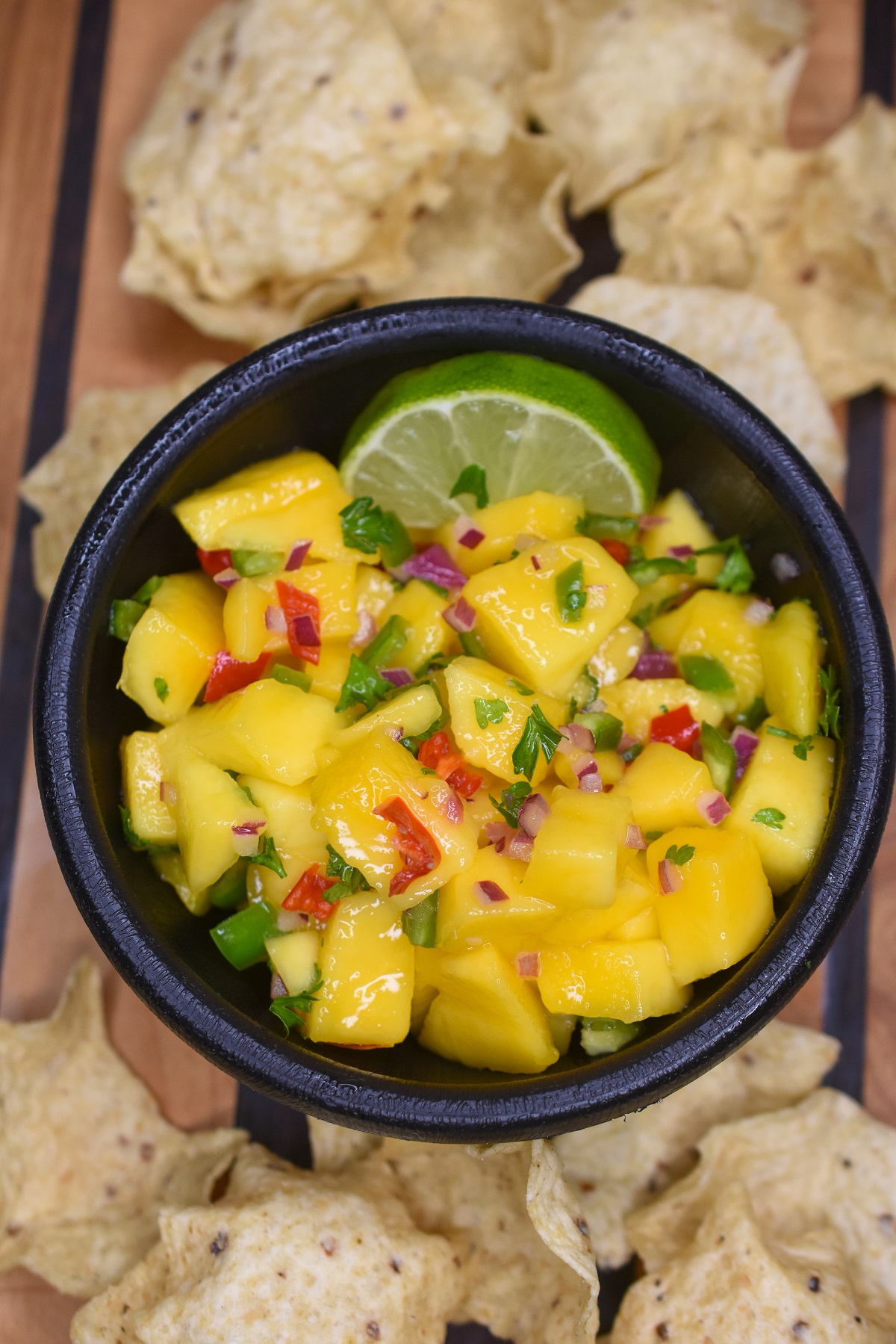 Mango Salsa recipe shown in a black bowl surrounded by tortilla chips garnished with a lime wedge.