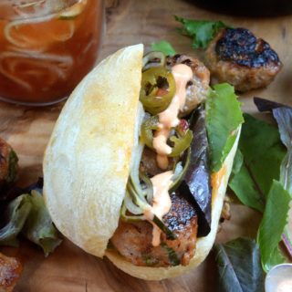 Pork Meatball Bahm Mi Sandwich. Shown mini meatballs on a crusty roll with greens pickled cucumber spirals, pickled jalapeno and sriracha mayo