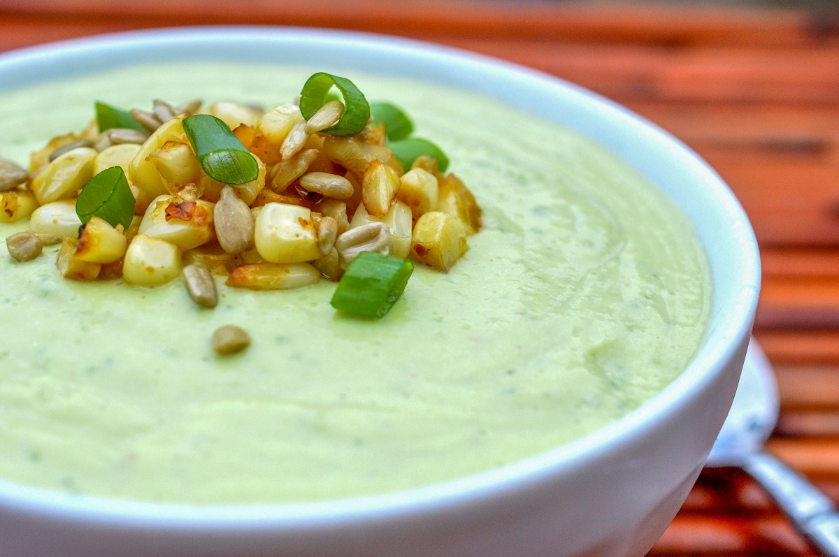 Avocado soup shown in white bowl topped with roasted corn kernels, sesame seeds and scallions. Green soup.