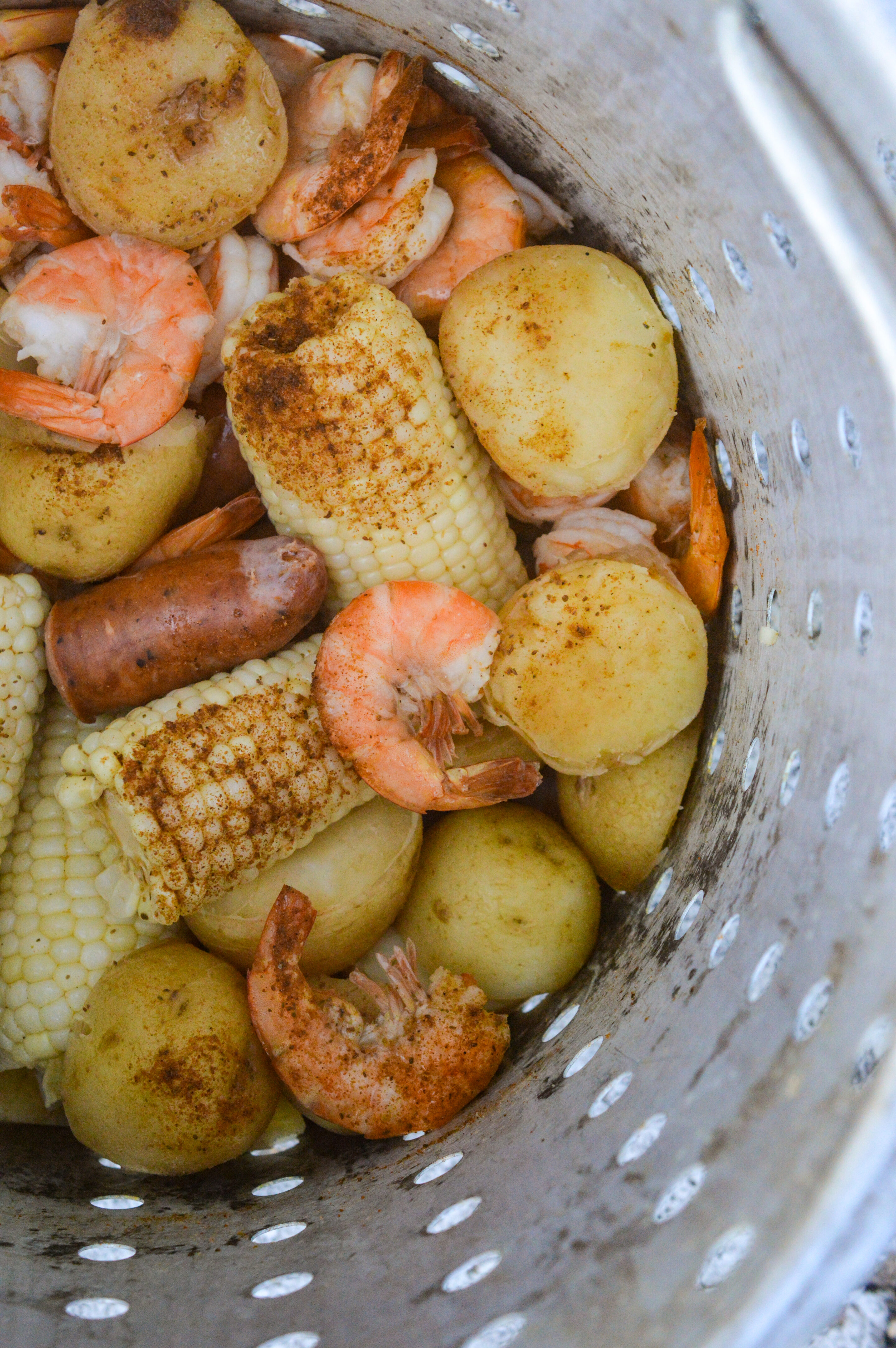 How to make a Shrimp Boil, also called Low Country Boil and Frogmore Stew. Make indoors or outdoors a delicious and fun way to feed a crowd! Shown a steamer pot full of corn segments, potatoes, shrimp in shells and andouille sausage with creole seasoning.