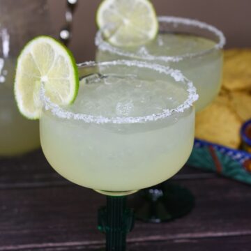 Classic Margaritas by the pitcher. Pitcer Margarita recipe. Shown pitche of margaritas in background. 2 cactus margarita glasses with salt rims and a lime filled with margarita mixture and ice.