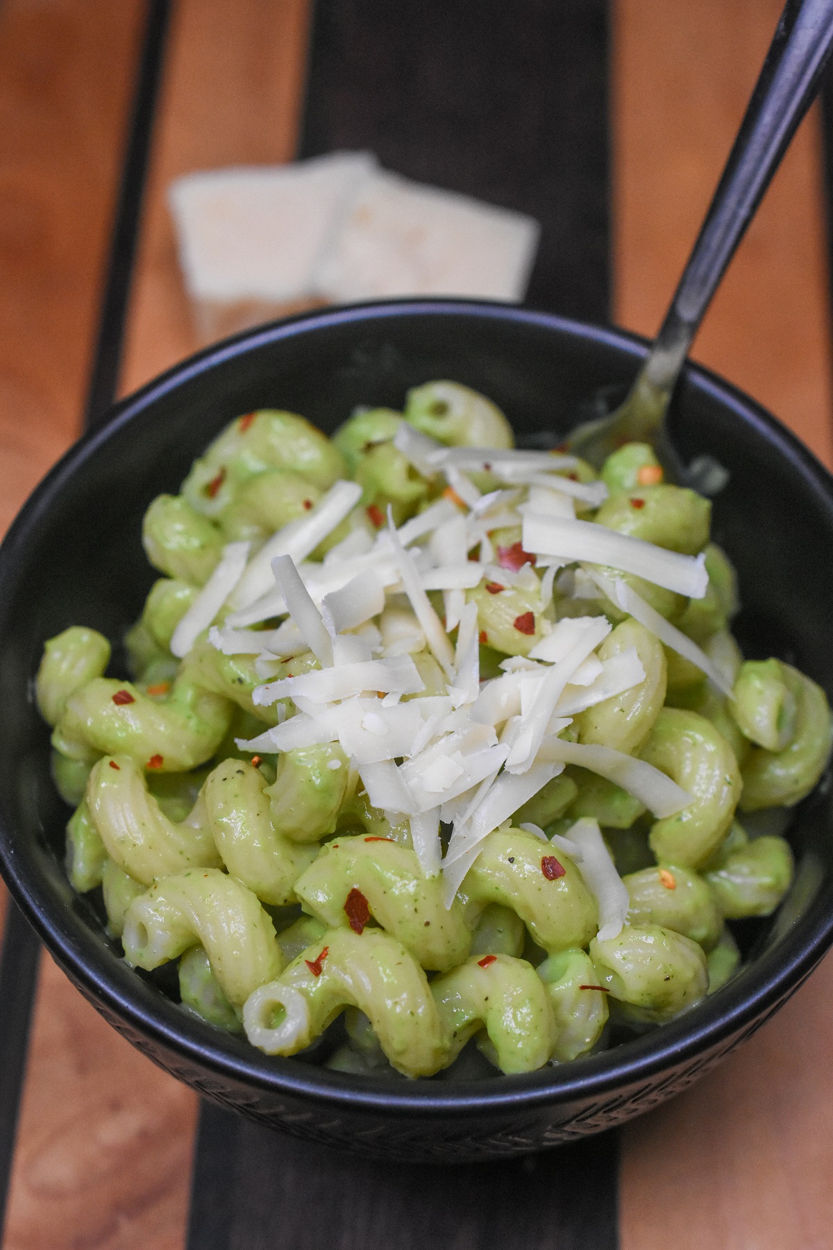 Cavatappi Pasta noodles in Avocado Sauce shown in a black bowl. Garnished with fresh shredded Parmesan cheese and red pepper flakes. Green Pasta.