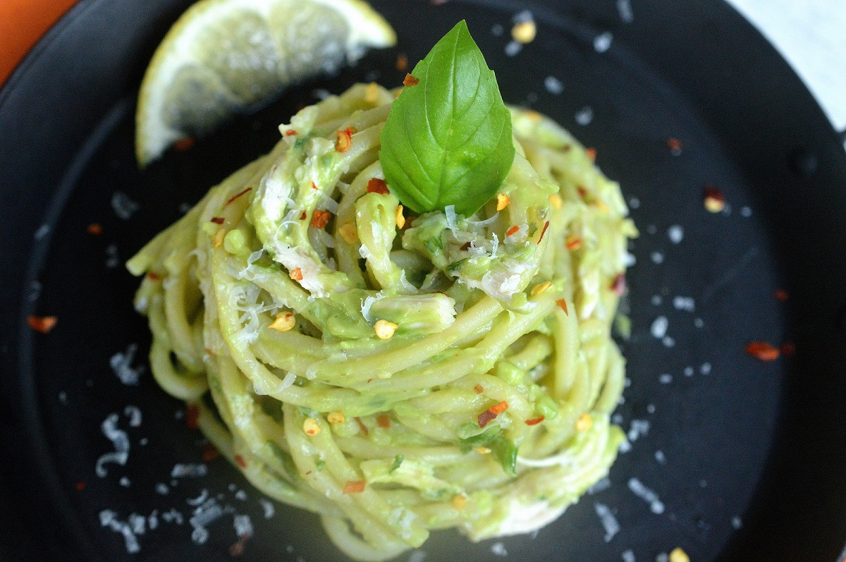 Green Pasta shown on black plate. Pasta with avocado pasta sauce, garnished with a lemon wedge, basil leaf and red pepper flakes,