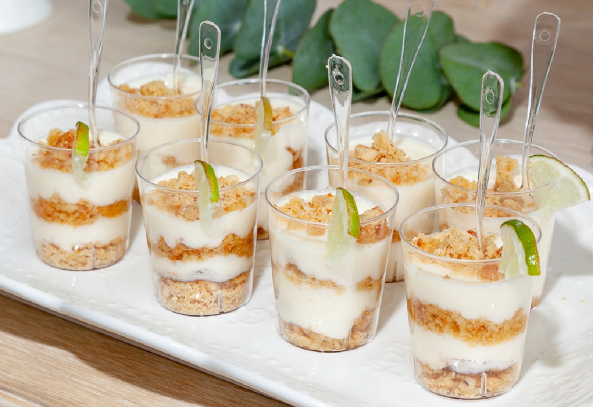 No bake Key Lime Pie Shooters shown on a white tray plate in 8 disposable shot glasses wit spoons. Graham cracker layers, key lime pie layers and a line wedge.