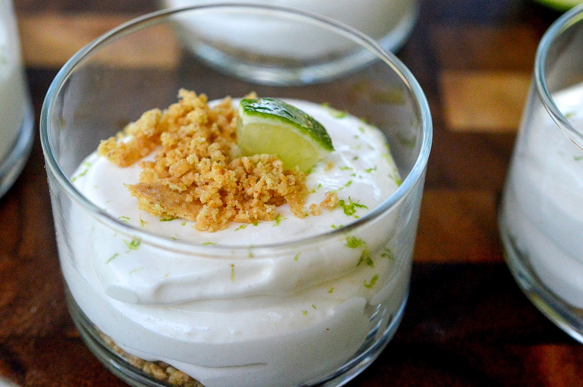 No Bake Key Lime Pie Dessert. shown in clear glass shooter cups. Graham cracker crumbs on bottom, key lime pie filling topped with more crumbs, lime zest and a lime wedge.