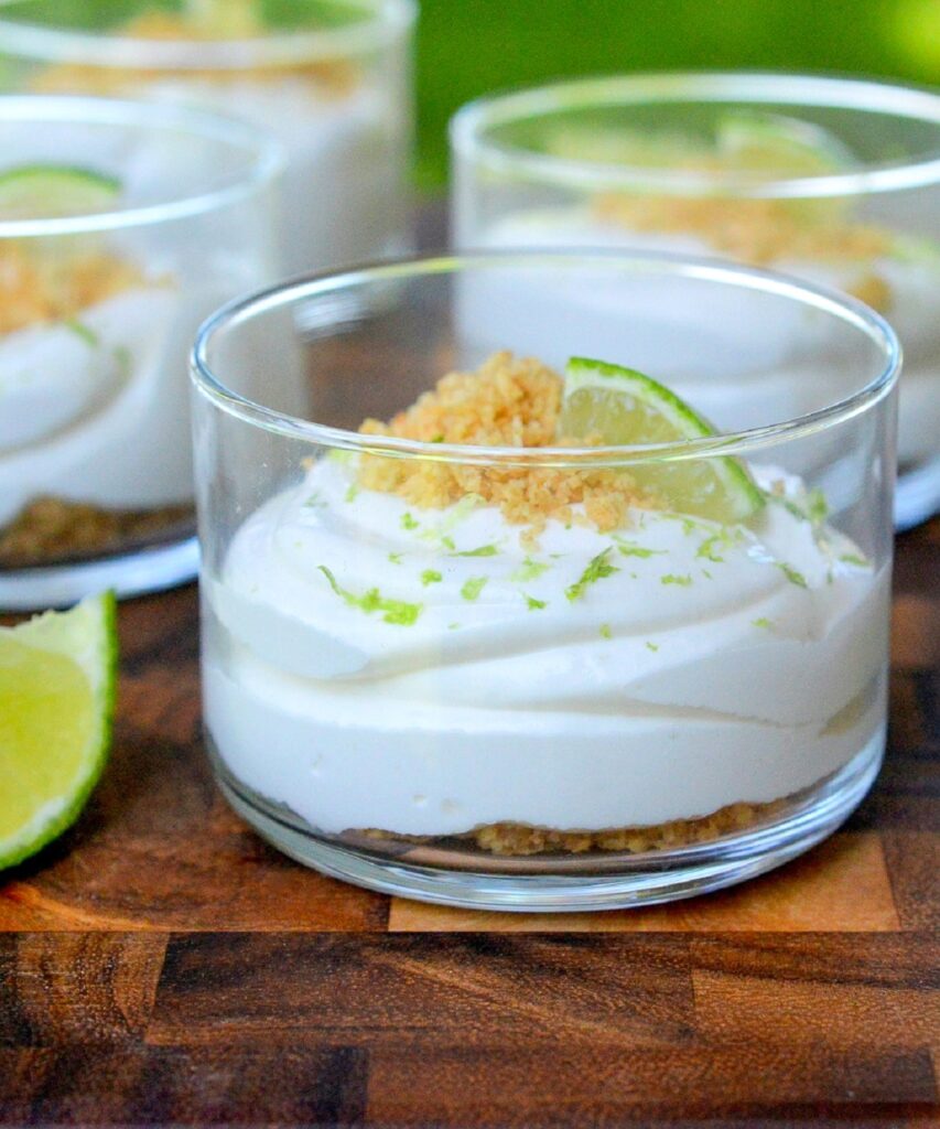 No Bake Key Lime Pie Dessert. shown in clear glass shooter cups. Graham cracker crumbs on bottom, key lime pie filling topped with more crumbs, lime zest and a lime wedge.
