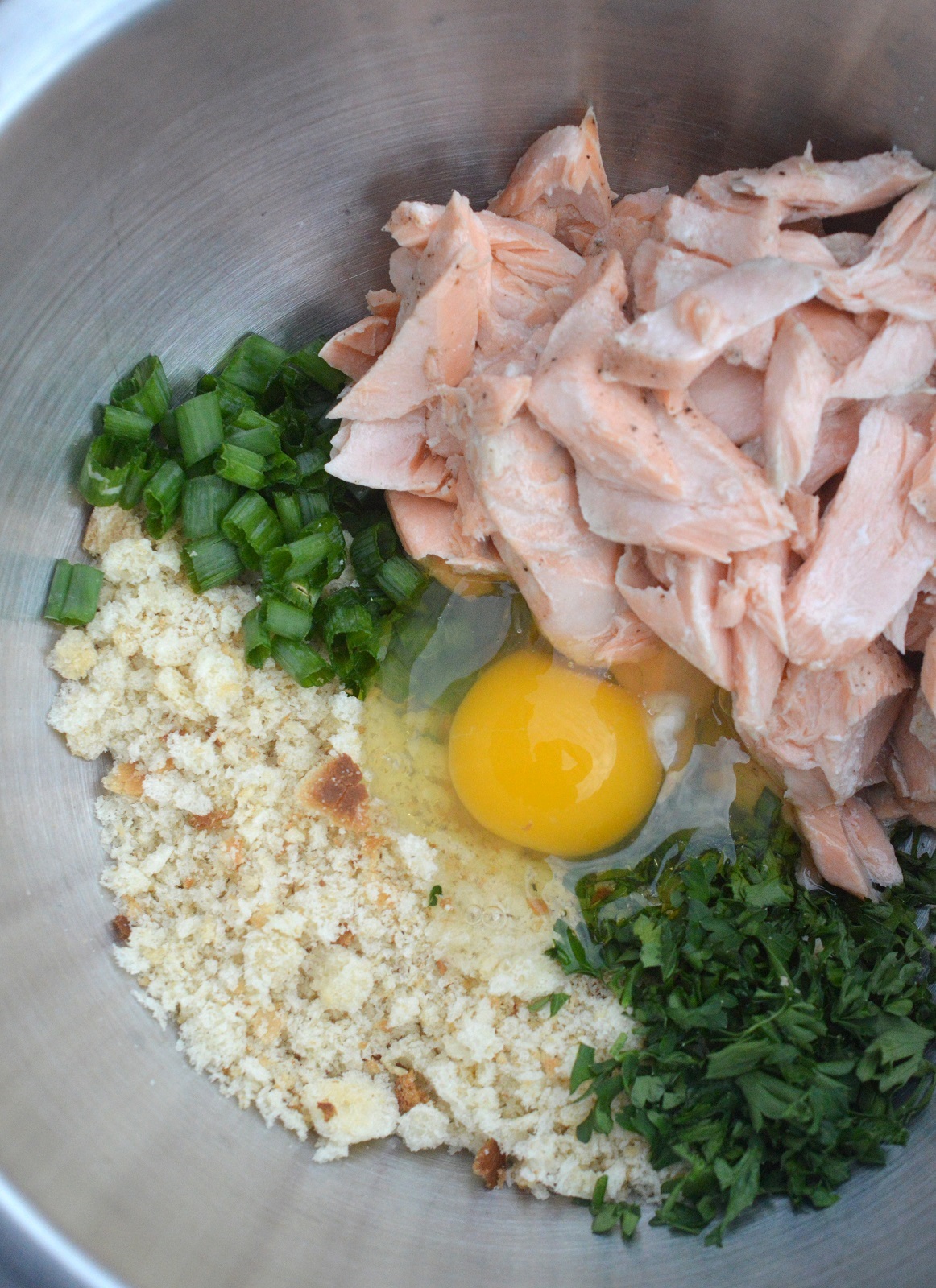 Image of ingredients in a bowl used to make salmon cakes. Flaked salmon, scallions, breadcrumbs, an egg and parsley