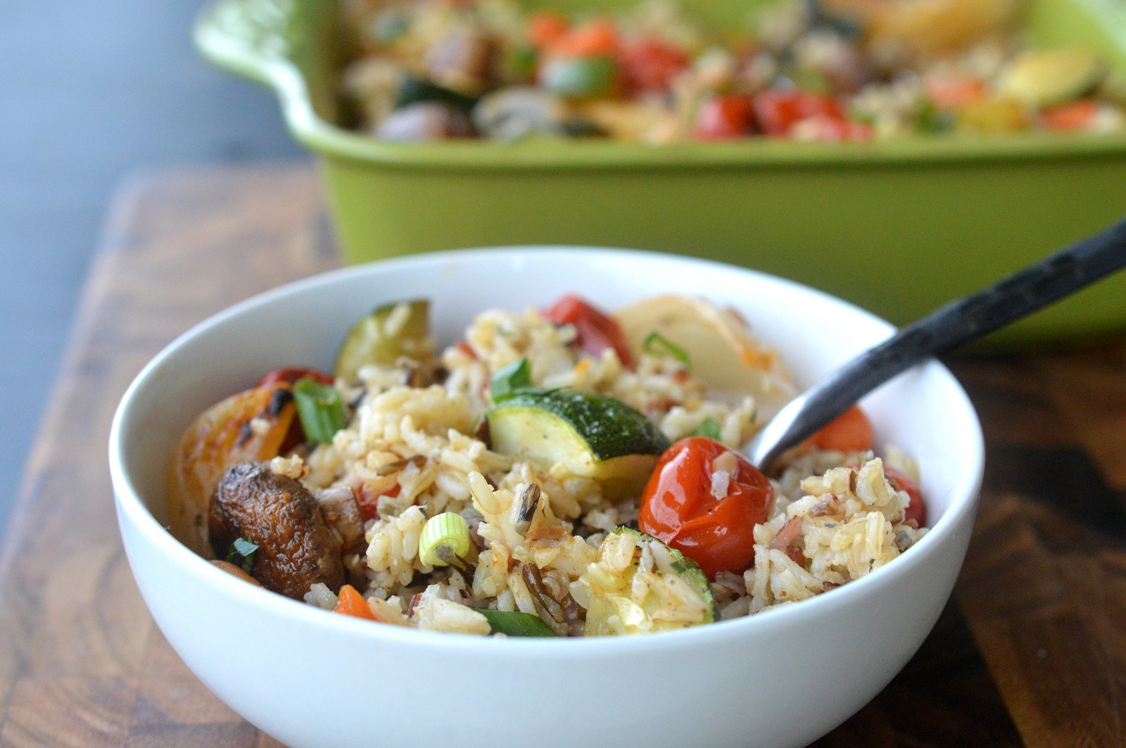 Roasted Vegetable Rice Recipe Shown Vegetable Rice in a white bowl with a fork. Behind it is a green casserole dish with Roasted Vegetables like tomatoes, zucchini and mushrooms and Rice.