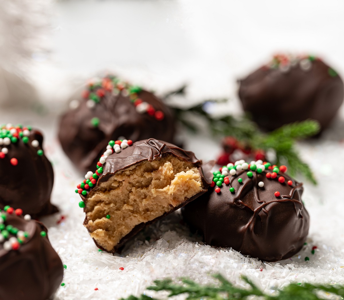 Chocolate Peanut Butter Balls also called Peanut Butter Truffles. Peanut Butter Balls dipped in chocolate decorated witrh holiday sprinkles. Shown on a Christmas placemat with one candy cut open. 