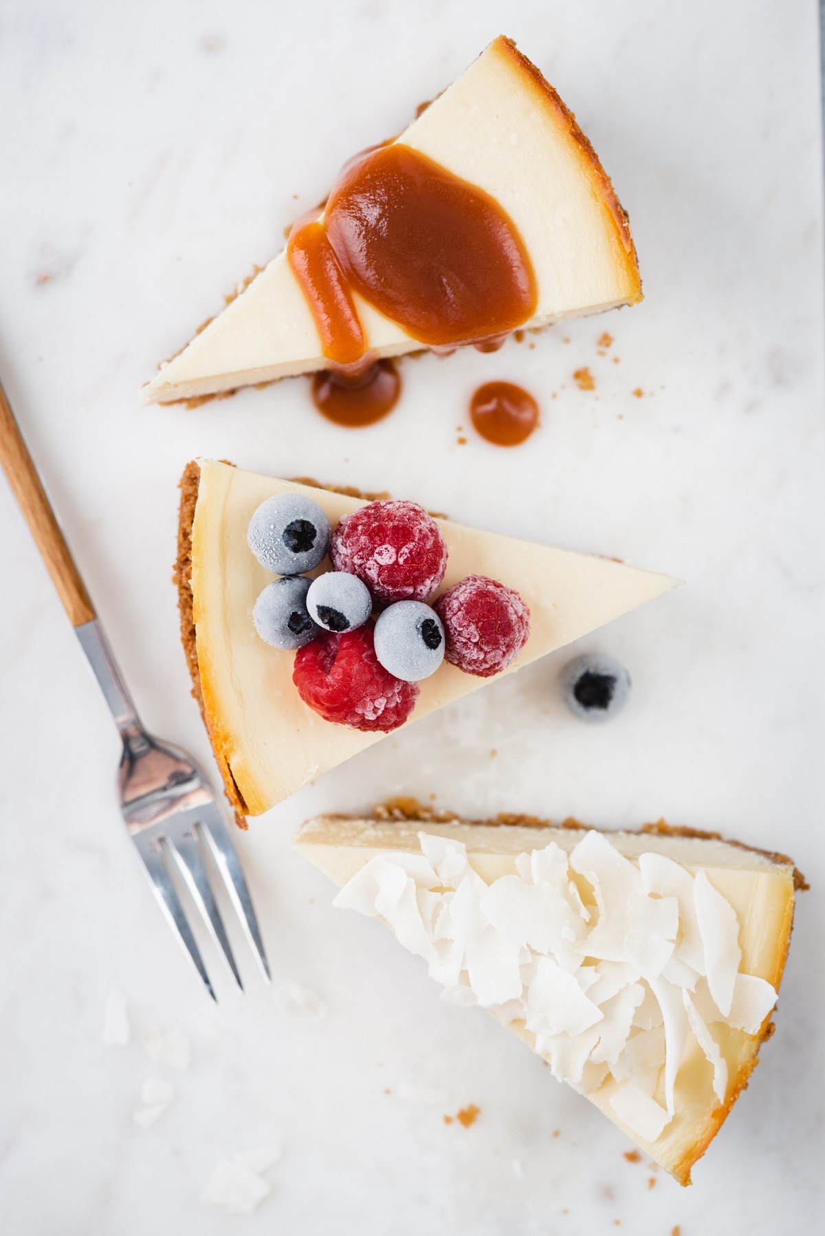 3 slices of cheesecake. one topped with white chocolate shavings, one topped with fresh fruit and one topped with caramel