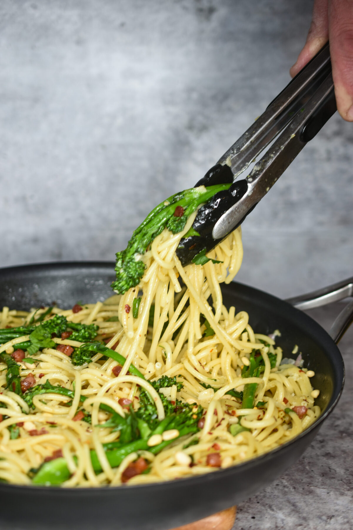 Pasta with Pancetta and broccoli rabe with pine nuts shown in a skillet with tongs picking up some of the pasta.