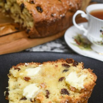 This really is the best recipe for Irish Soda Bread. It's a sweeter more cake-like soda bread that always gets rave reviews. Shown a slice of Irish Soda Bread with raisins with butter on a plate with a cup of tea. Full loaf in background.