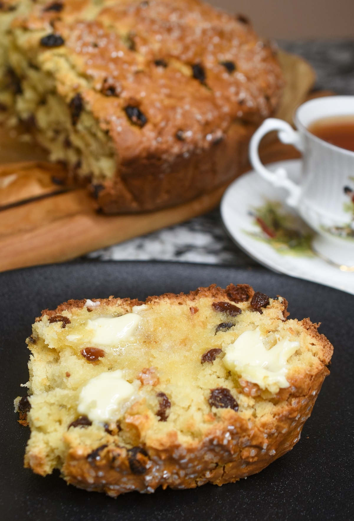 This really is the best recipe for Irish Soda Bread. It's a sweeter more cake-like soda bread that always gets rave reviews. Shown a slice of Irish Soda Bread with raisins with butter on a black plate. A cup of tea on Royal Tara China in background as well as a cut open loaf of soda bread on a wood cutting board.