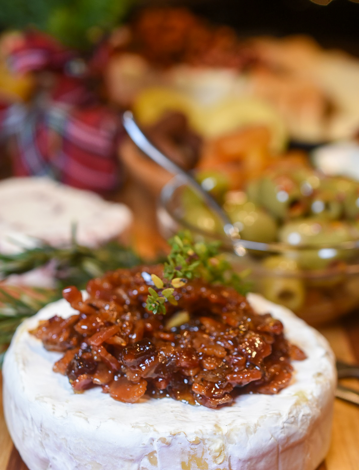 Bacon Jam topped Brie Cheese wheel on a charcuterie board. with olives in background.