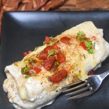 one white chicken enchilada on black plate with fork garnished with chopped seasoned tomatoes and herbs