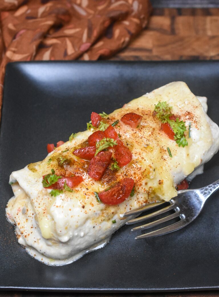 one white chicken enchilada on black plate with fork garnished with chopped seasoned tomatoes and herbs