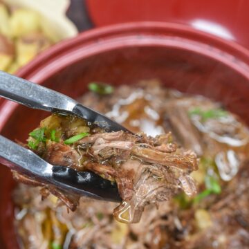 Oven roasted chuck roast pulled apart shown in Dutch oven with tongs holding shredded chuck roast