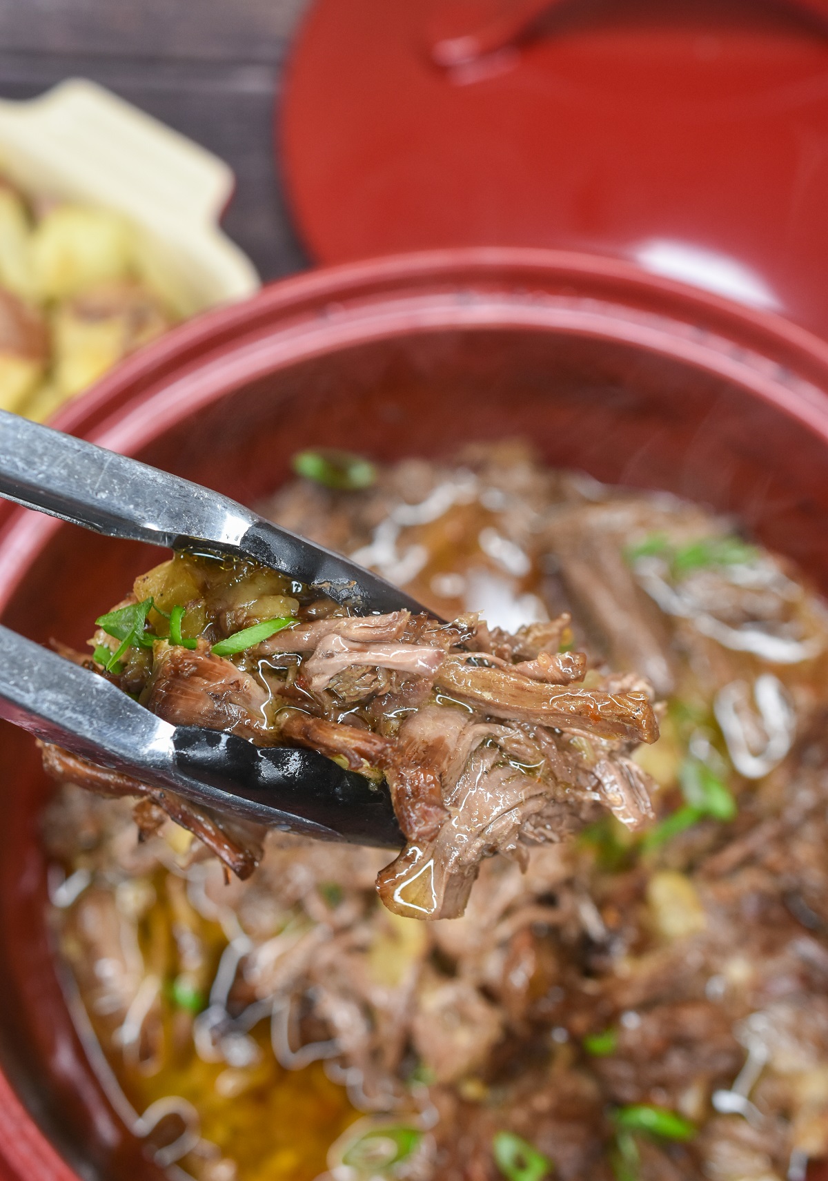 Oven roasted chuck roast pulled apart shown in Dutch oven with tongs holding shredded chuck roast