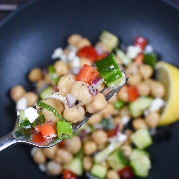 Dense Bean Salad recpe shown in a blck bowl with some bean salad on a fork. You can see chickpeas, cucumbers, red onion, parsley, feta cheese & red pepper.