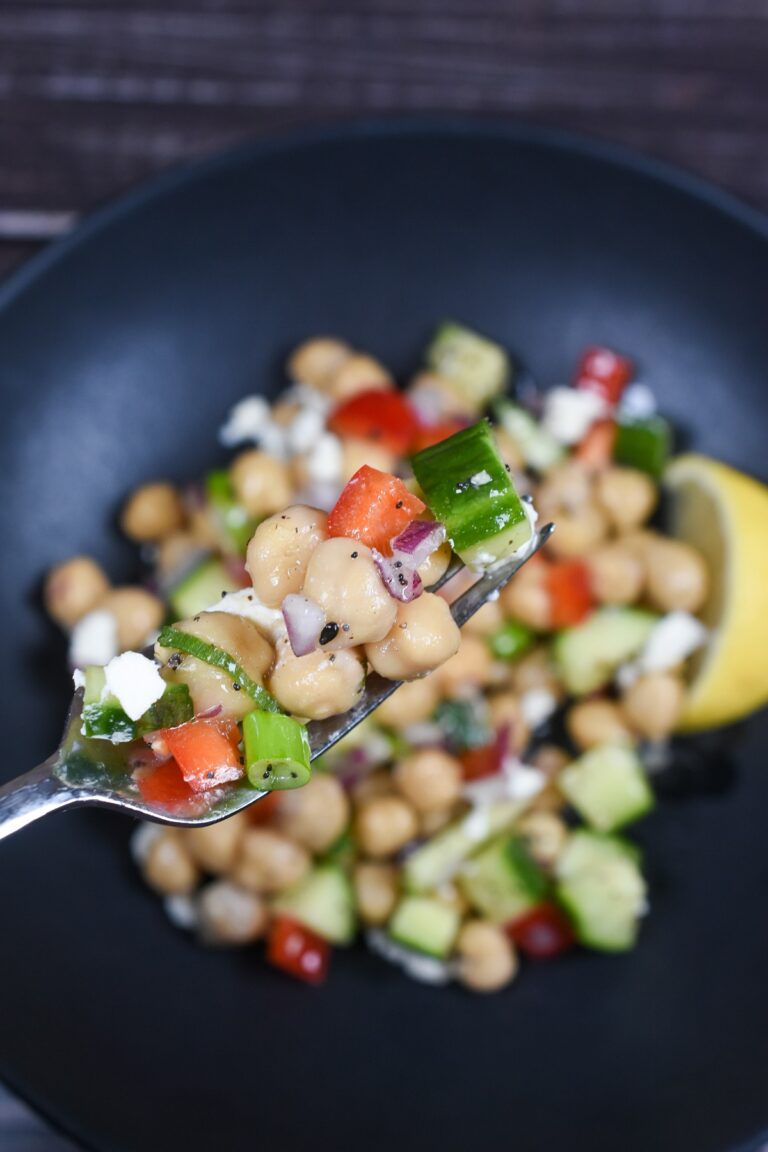 Dense Bean Salad recpe shown in a blck bowl with some bean salad on a fork. You can see chickpeas, cucumbers, red onion, parsley, feta cheese & red pepper.