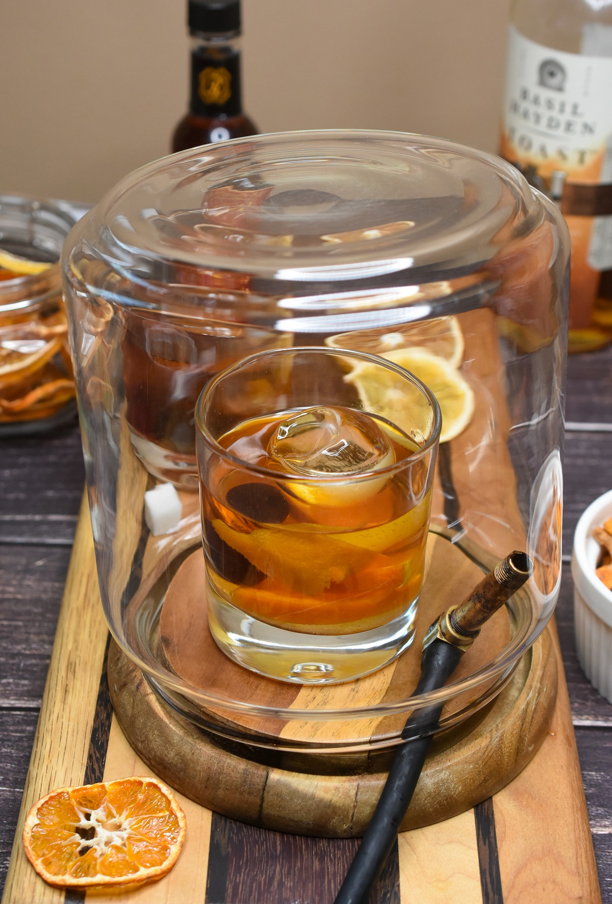 Making a SMoked Old Fashioned. Smoker glass overtop old fashioned cocktail with smoker tube under glass. Dried orange slices shown in jar and bottle of bourbon in background.