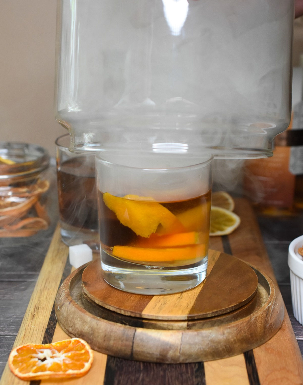 Making a Smoked Old Fashioned Drink. Shown smoking and old fashioned. A rocks glass with smoker filled with smoke being lifted, dried oranges in background. Bourbon Old Fashioned