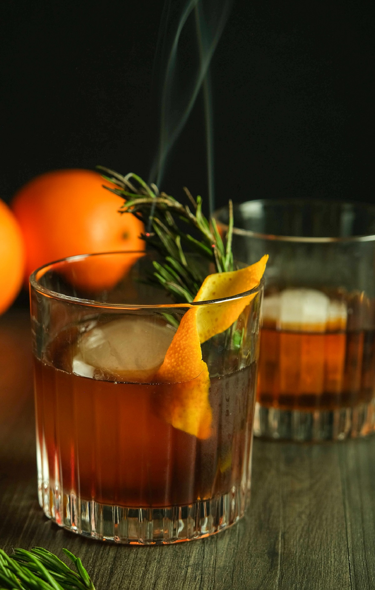 Old Fashioned Drink shown with fresh Rosemary Sprigs smoking in the drink. Orange peel twish garnish, oranges in background. How to make Smoked Old Fashioned.