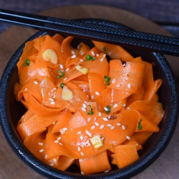 Korean Carrot Salad also called Morchovka is delicious and easy to make. Shown thin ribbons of carrot in a black bowl with black chopsticks garnished with sesame seeds and parsley.