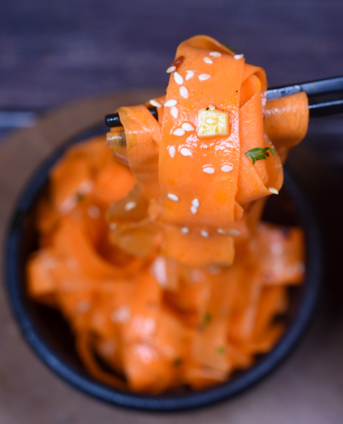 Korean Carrot Salad also called Morchovka is delicious and easy to make. Shown thin ribbons of carrot in a black bowl with black chopsticks lifting some of the carrot salad up garnished with sesame seeds and parsley.