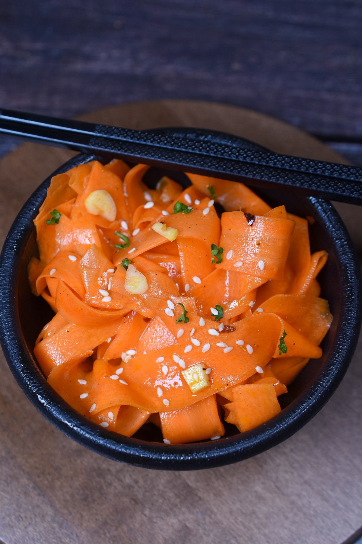 Korean Carrot Salad also called Morkovcha is delicious and easy to make. Shown thin ribbons of carrot in a black bowl with black chopsticks garnished with sesame seeds and parsley.