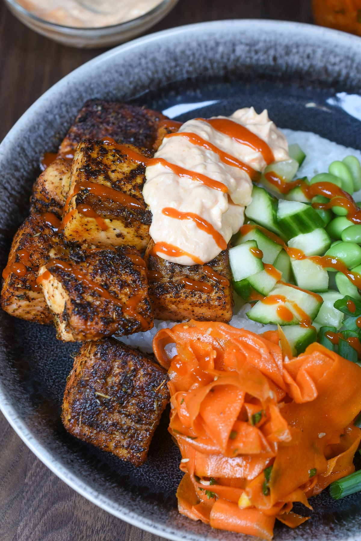 Blackened Salmon Rice Bowl such a quick, easy and satisfying meal.. Shown blackened salmon bites over rice with edamame, Korean carrot salad, scallions and cucumbers. with Sriracha mayo