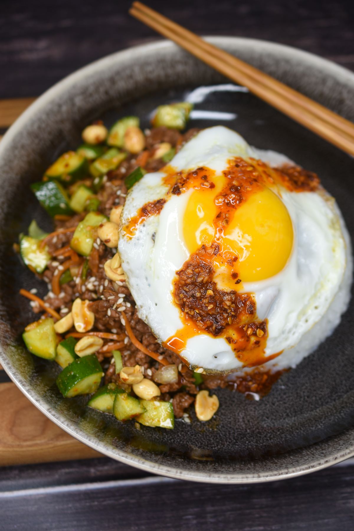 Ground Beef Stir Fry shown plated in clack bowl with sticky rice, spicy cucumbers, a fried egg with a drizzle of chili crisp and chopsticks