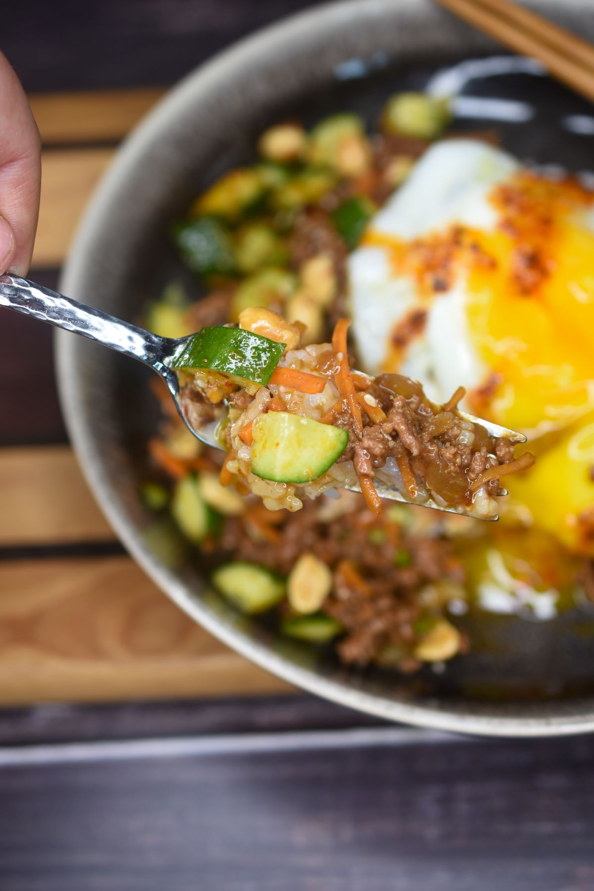 Ground Beef Stir Fry with Asian flavors topped with a fried egg, shown a forkful with ground beef, cucumber salad and rice overtop a bowlful with runny yolk