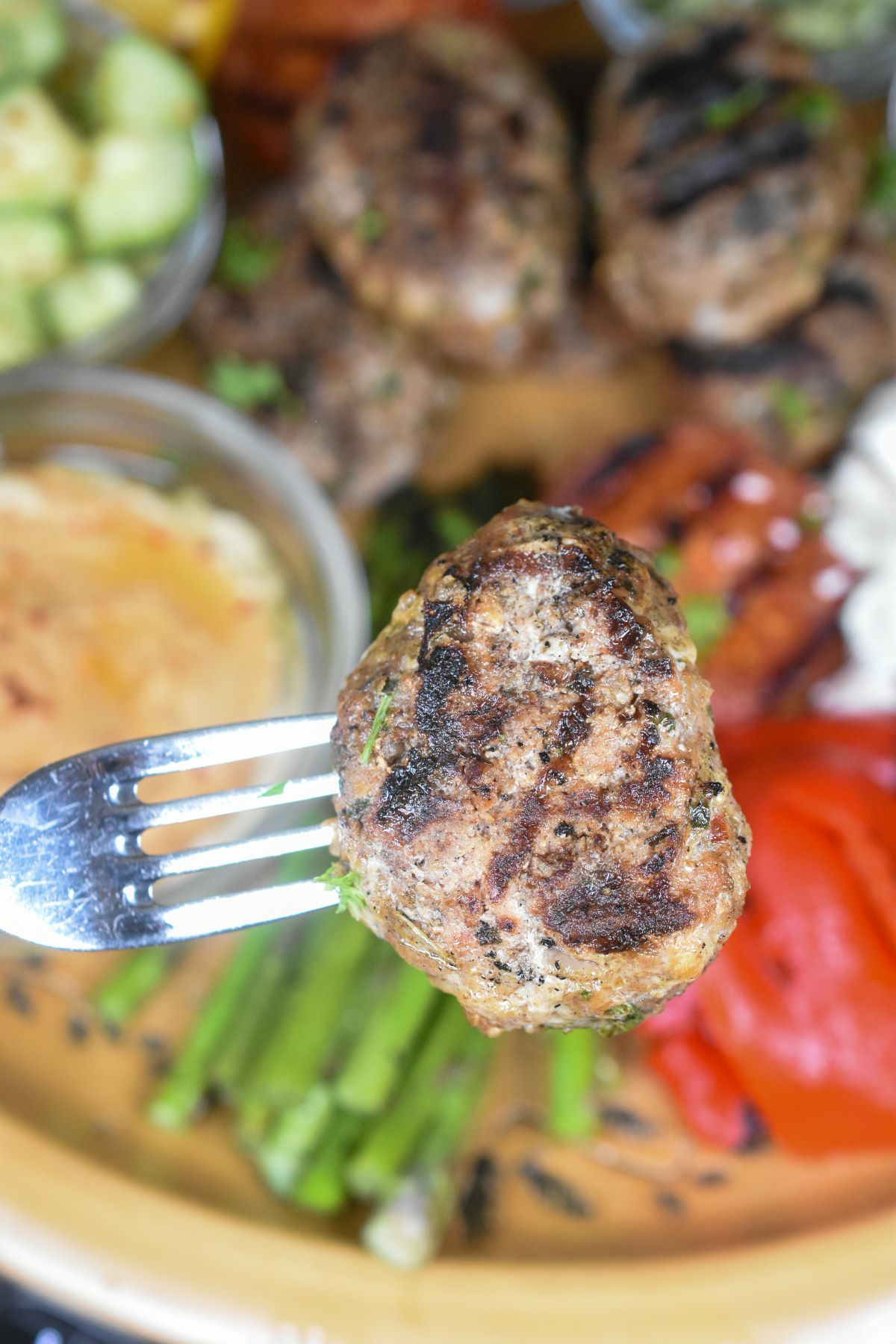 Grilled Mediterranean Platter with grilled kofta up close on a fork. Background of grilled asparagus, tomato halves, hummus and more.