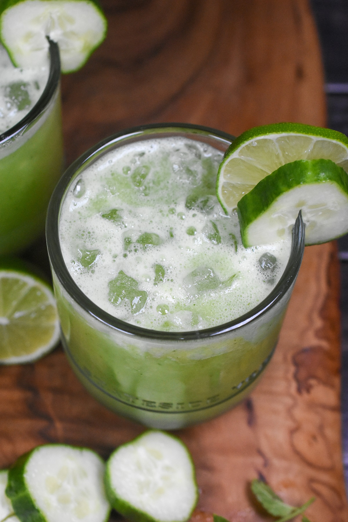 Cucumber Agua Fresca Recipe. Super refreshing cucumber drink recipe. Aqua De Pepino is a delicious and easy way to stay hydrated. . Shown green juice in a clear glass with ice. Sliced cucumbers and mint in background.