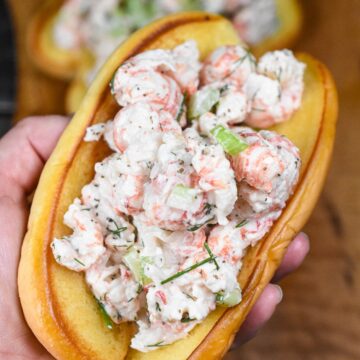 If you enjoy lobster rolls, you'll like these Langostino Lobster Rolls. Similar in taste and texture without the cost or the extra work. Shown is a roasted buttered potato hot dog bun loaded with langostino lobster salad held in a hand. Salad is made with langostino, mayo, dill, celery, chives, lemon juice, salt and pepper.
