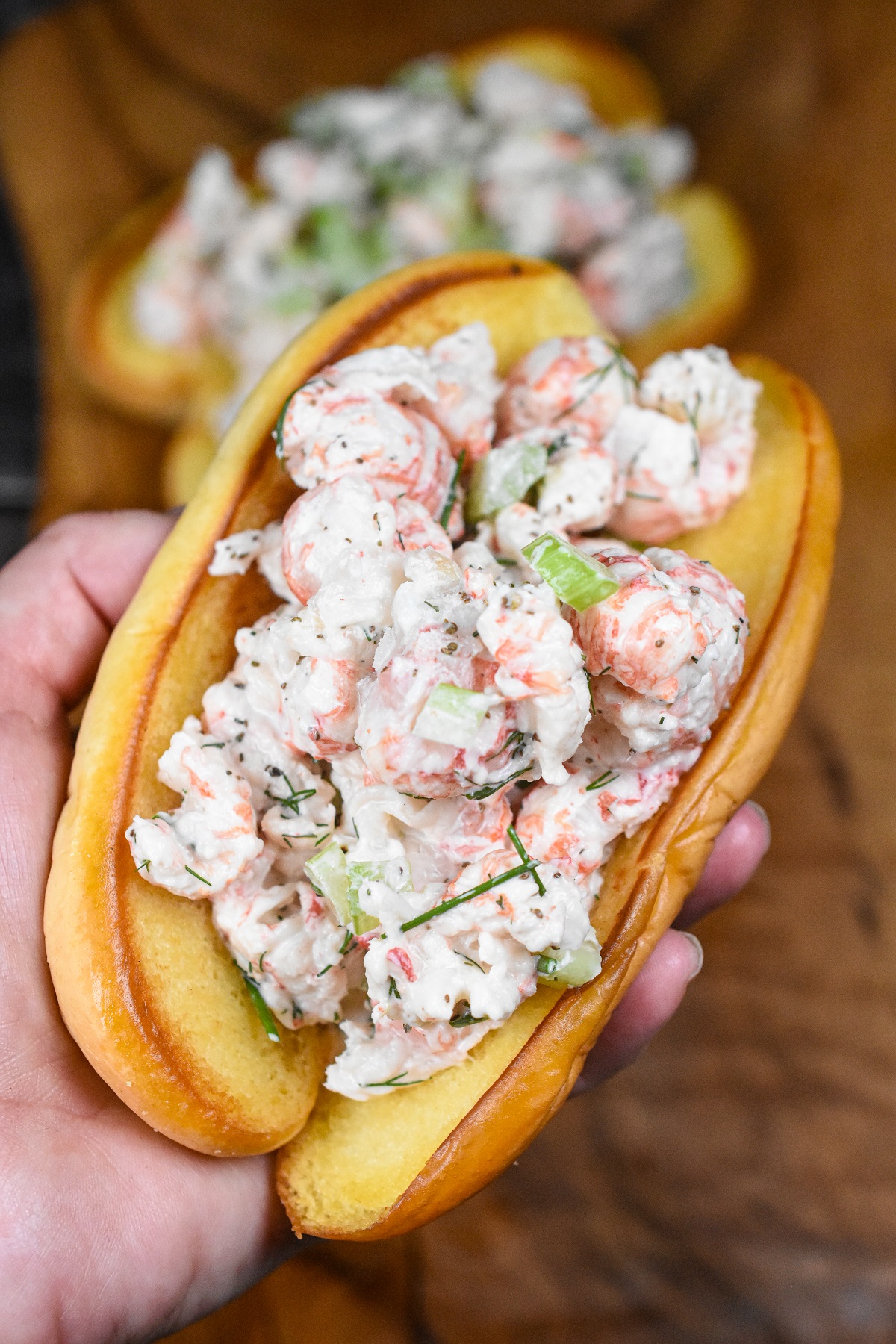 Langostino Lobster Rolls, shown langostino lobster salad piled on a buttered & toasted potato bun held in a hand.