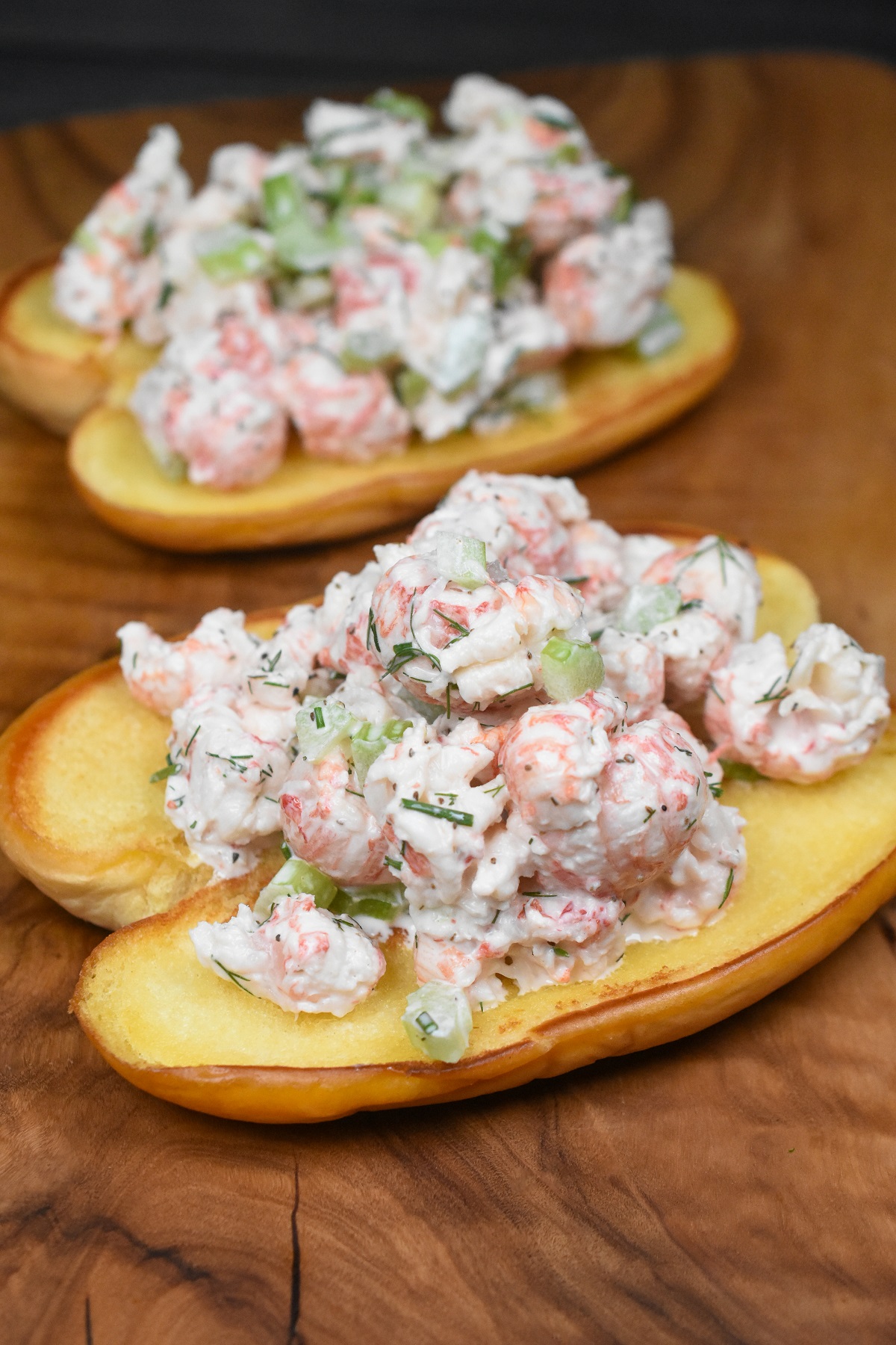 Langostino Lobster recipe, Shown, 2 langostino lobster rolls on a wood cutting board. Toasted and buttered hot dog rolls piled high with langostino lobster salad.