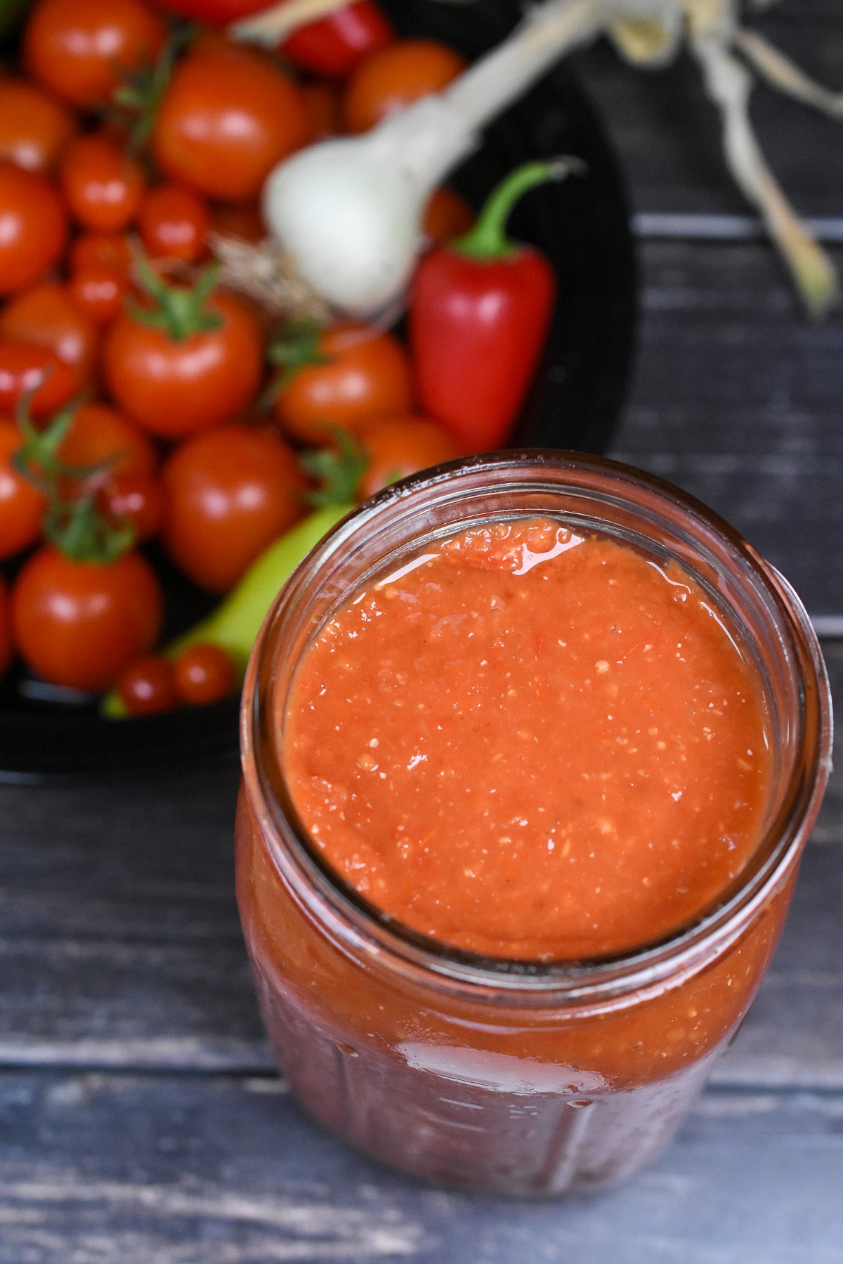 Make an amazing oven Roasted Tomato Sauce in 30 minutes. Great for pasta dishes, stews, soups and more. Freezes very well! Shown a quart jar full of roasted tomato sauce with a bowl of garden tomatoes, peppers, onion and garlic.