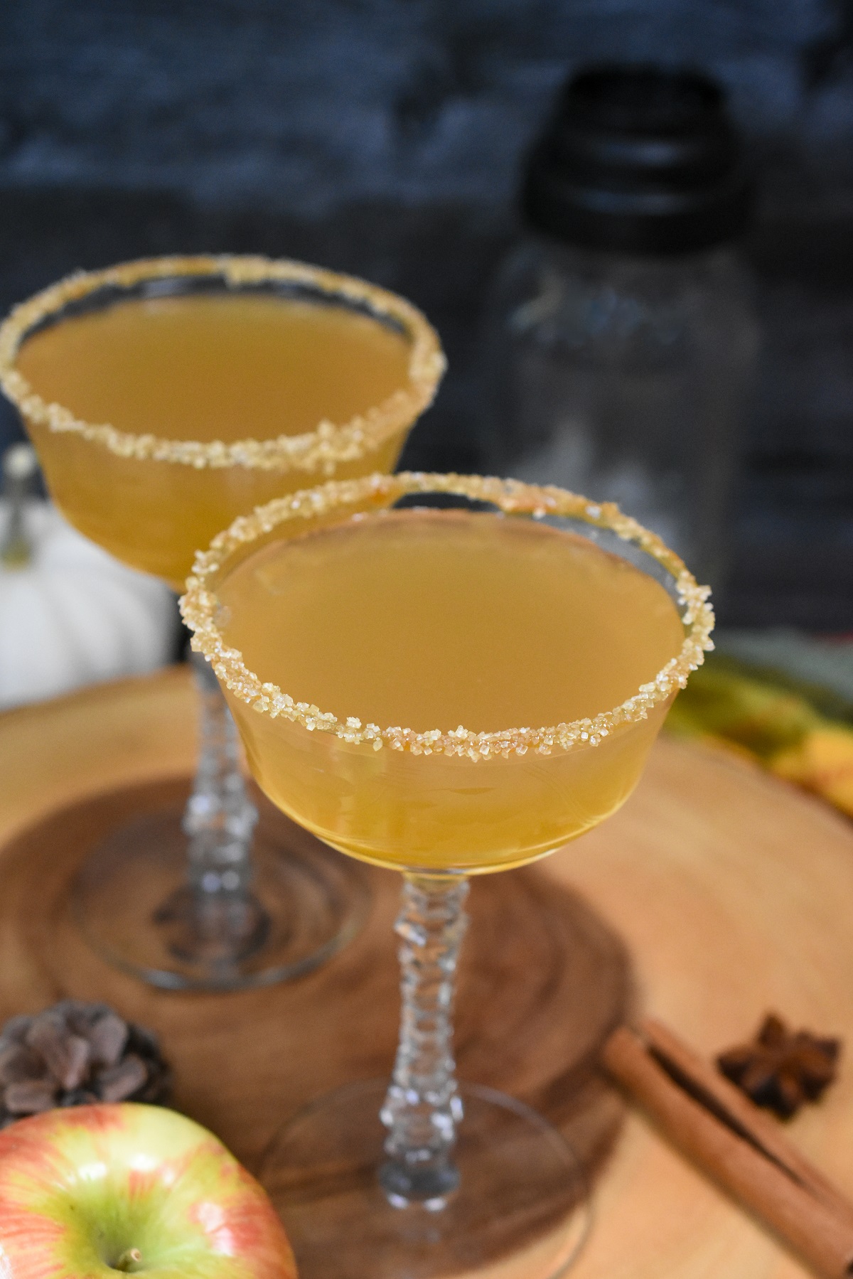 Apple Cider Martini recipe, shown apple cider martini in two vintage coupe glasses rimmed with caramel and sugar on a wood cutting board with cinnamon sticks, an apple and pine cone scattered.