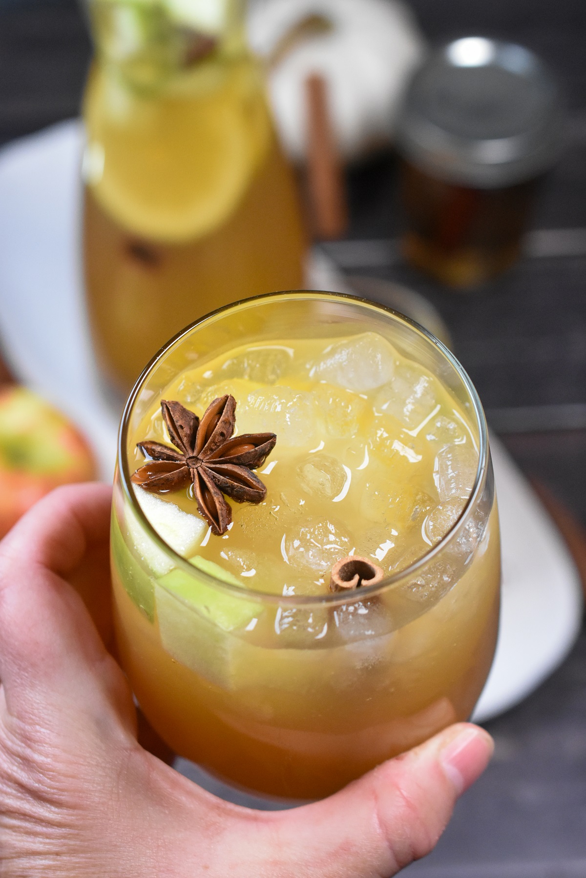 Fall cocktail made using Cinnamon Syrup. Apple Cider Sangria garnished with cinnamon stick, chopped apples and a star anise. Glass held by a hand with ice and liquid.. 