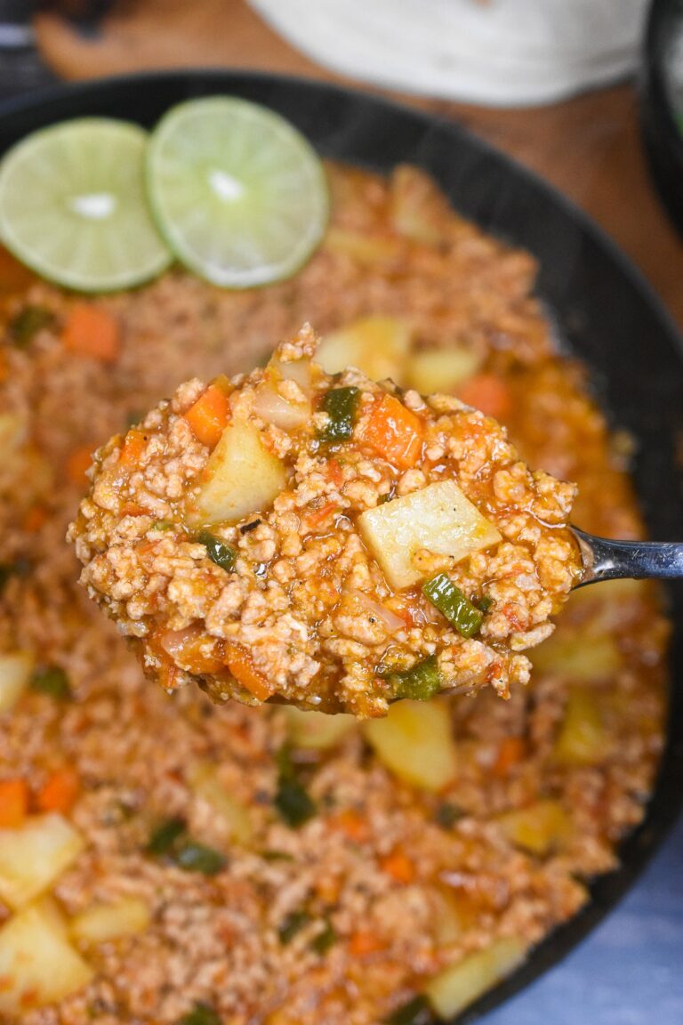 Picadillo shown cooked in a large cast iron pan, lime slices as garnish and a silver spook lifting some of the Picadillo up from the pan.
